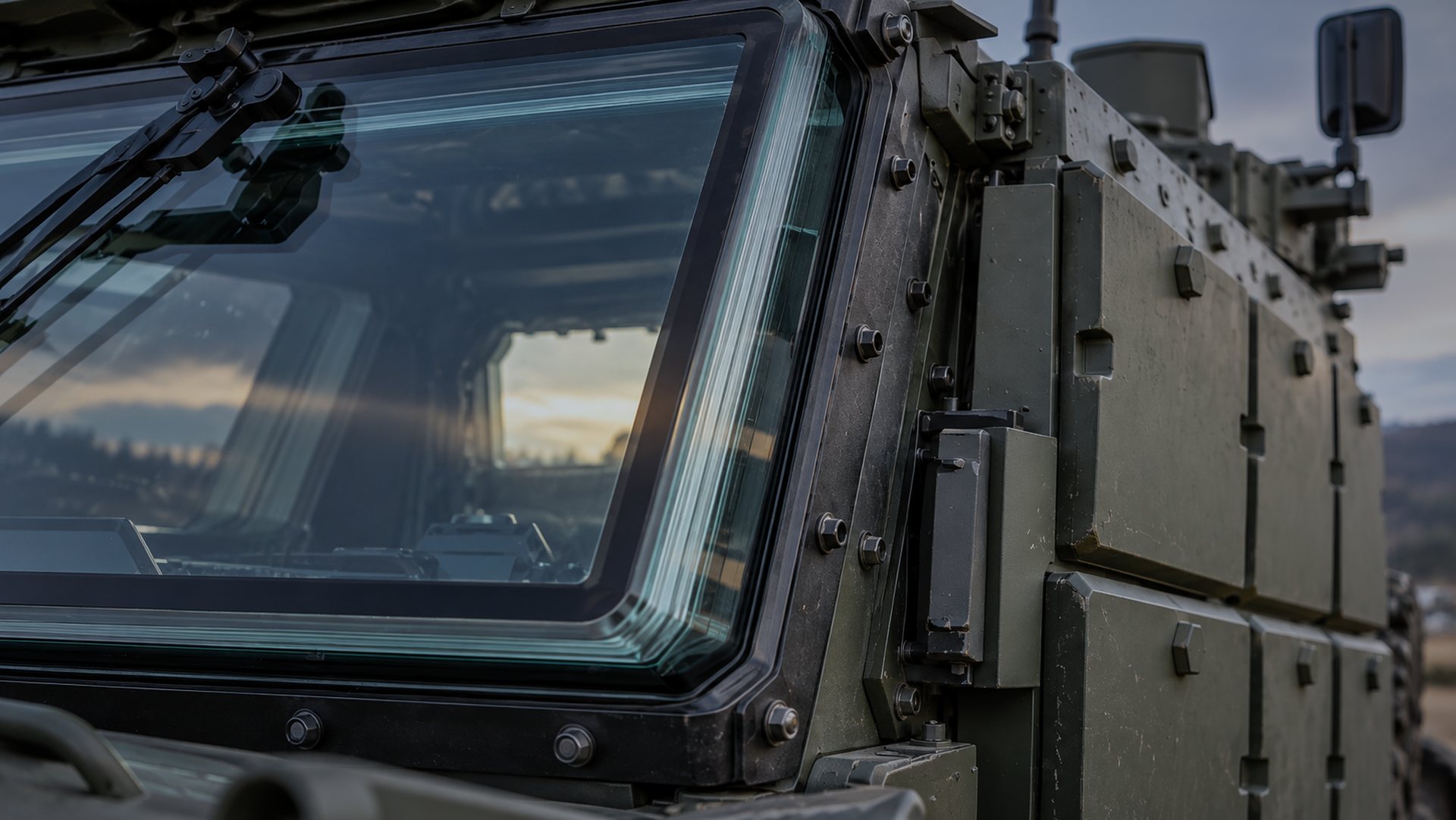 Close view of a protected vehicle's transparent armour windscreen and its bolted frame, with modular protection panels along the hull.